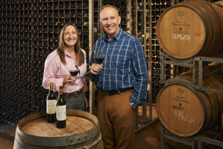 Clare and Keith Mugford standing in the cellar holding glasses of wine