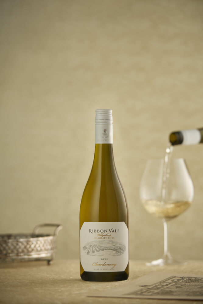 02_20230131_MOSS WOOD_Creatives_Chardonnay 2022_Vertical_POURING VERSION A bottle of Ribbon Vale 2022 Chardonnay with a glass being poured in the background